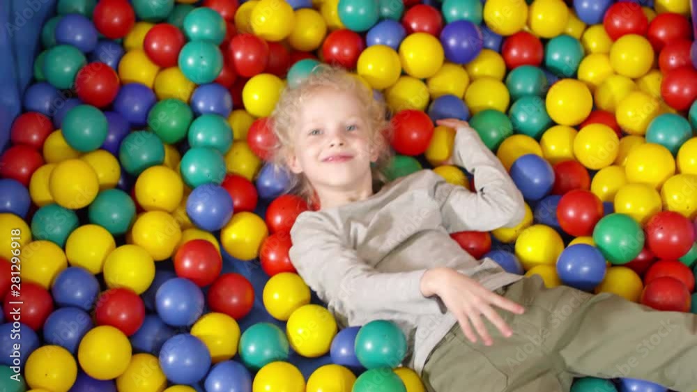 Handheld top view shot of adorable little girl lying in ball pit and throwing colorful balls at camera Handheld top view shot of adorable little girl lying in ball pit and throwing colorful balls at camera