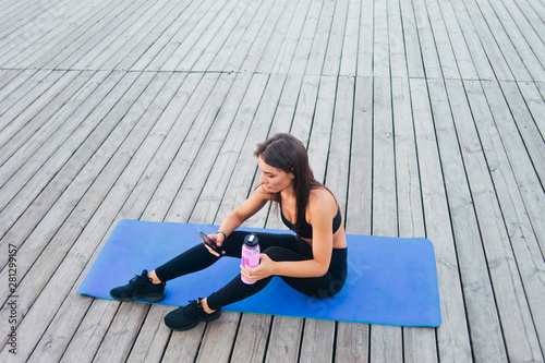Cheerful healthy fitness woman in sportswear sitting on  yoga mat and chatting in smartphone outdoor on wooden boards