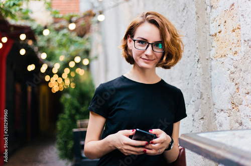 Young hipster girl in glasses with short haircuts use smartphone and sitting in outdoor street cafe photo