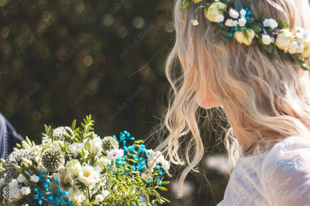 Naklejka premium Beautiful blonde young bride portrait with flower bouquet and wreath on her head in white wedding dress outdoor in summer