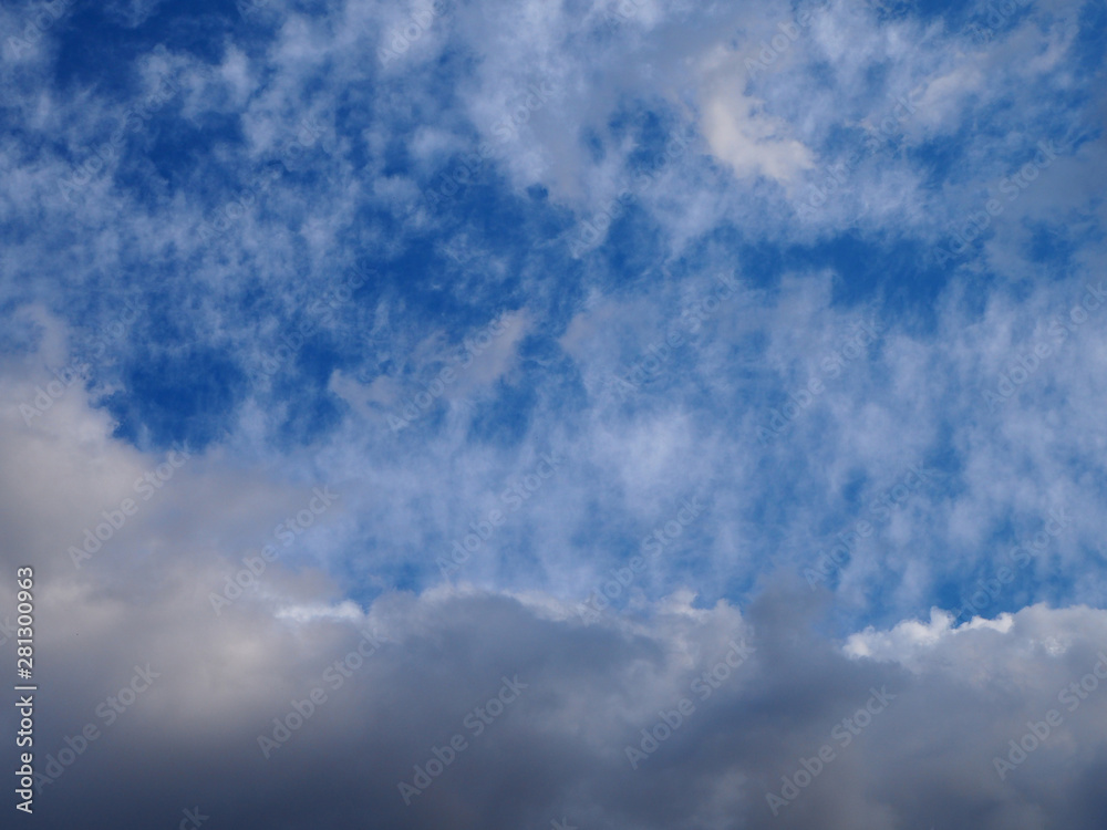 Fototapeta premium Landscape of beautiful blue sky covered with light thin fluffy white clouds and a thundercloud coming from below. Natural cloudy blue sky on day time abstract background.