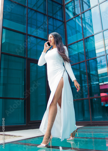 Fashion afro woman in a luxurious white dress talking on phone while walking against building with blue windows outdoor.