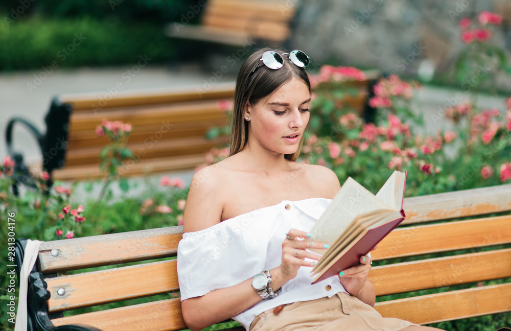 Obraz premium Young blonde woman reading a book with enthusiasm while sitting on a bench in a park