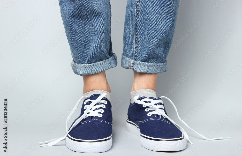 Fashion studio shot. Female legs in jeans and sneakers with untied laces on gray background. Fashion studio shot. Female legs in jeans and sneakers with untied laces on gray background.