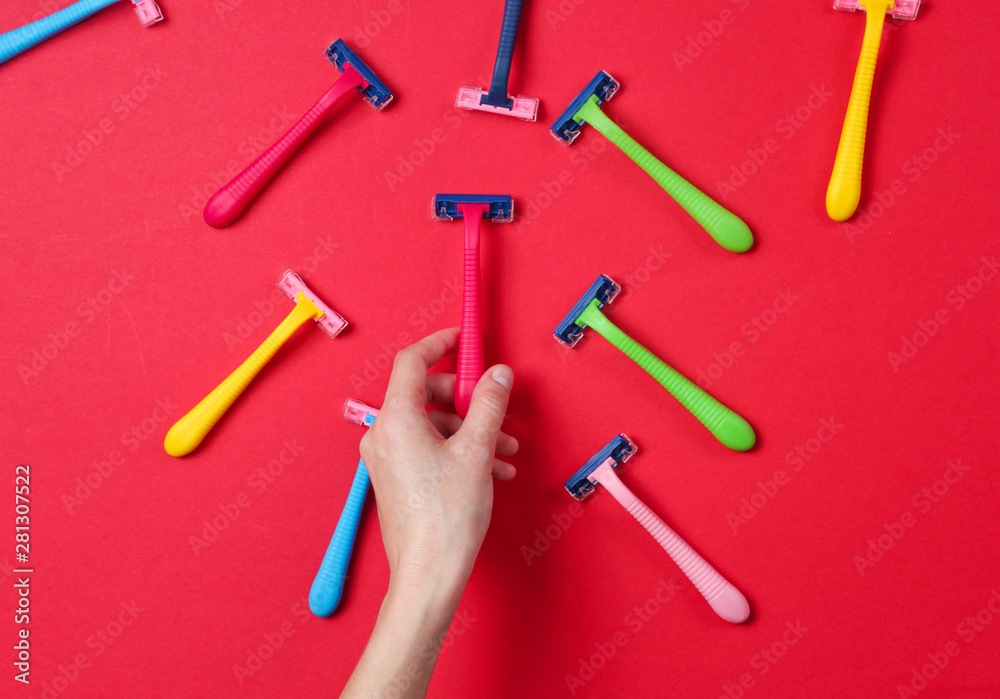 Female hand choosing green razor among a variety of colored epilator razors on red background. Top view. Minimalism beauty concept. Top view Female hand choosing green razor among a variety of colored epilator razors on red background. Top view. Minimalism beauty concept. Top view