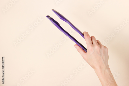 Female hand is holding kitchen tweezers on beige background. Top view