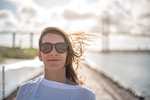 Woman wearing sunglasses and white light clothes looks forward, with a sunny tropical landscape behind.