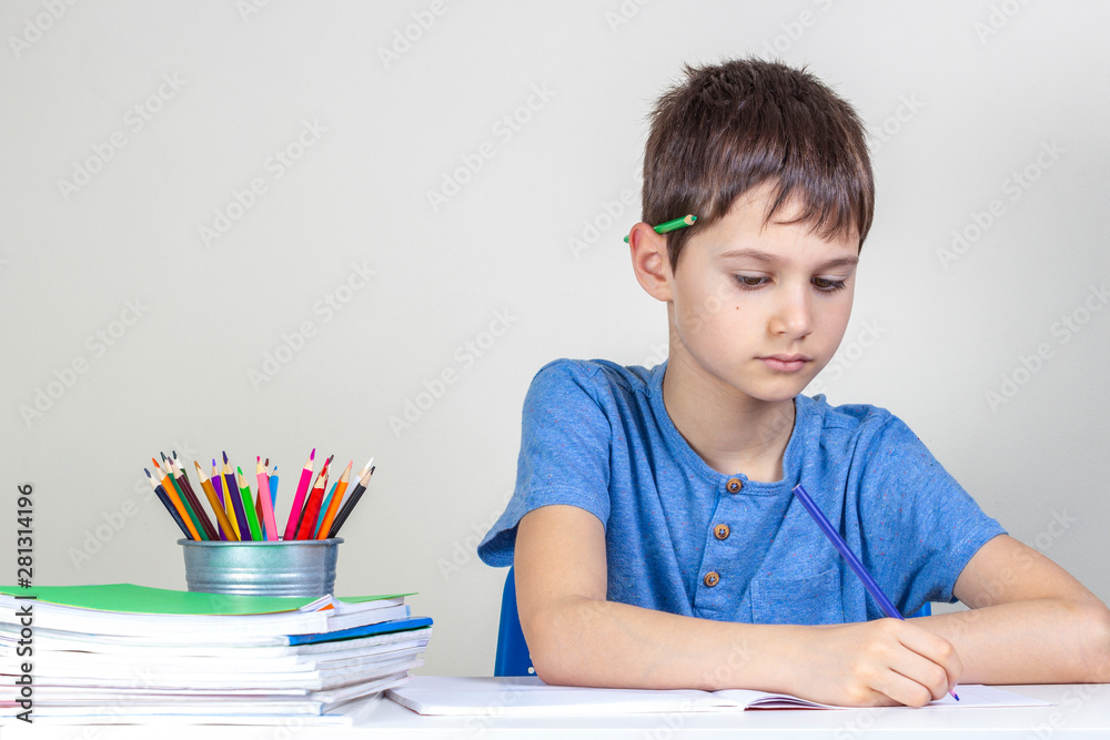 Kid doing homework at the table. Focused boy with pencil behind his ear writing with pencil Kid doing homework at the table. Focused boy with pencil behind his ear writing with pencil