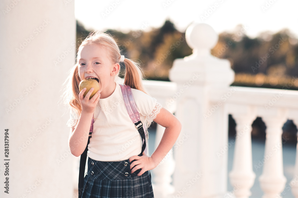 Funny child girl 5-6 year old biting green fresh apple outdoors. Wearing school uniform and backpack. Back to school. 1 September.