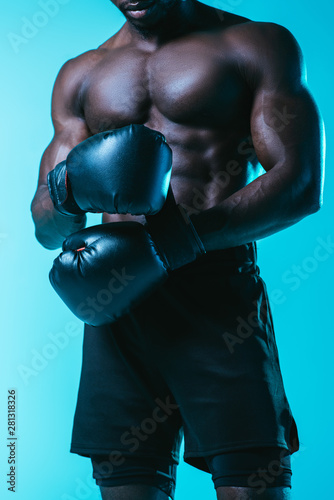 partial view of shirtless, muscular african american sportsman in boxing gloves on blue background