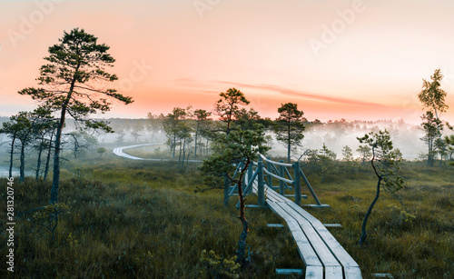 Obraz na płótnie Warmly colored sunrise over a foggy swamp. Aerial view of stunning landscape at peat bog at Kemeri National park in Latvia. Wooden trail leading along the lake surrounded by pounds and forest. w salonie