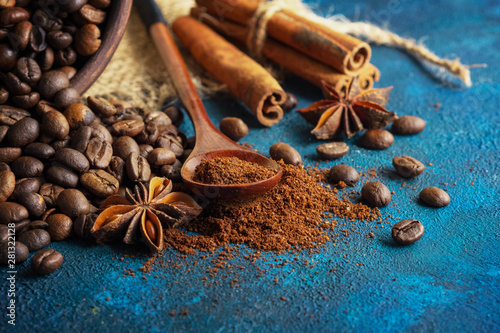 coffee grains scattered on a blue textural background, anise stars, cinnamon sticks and ground coffee in a wooden spoon.