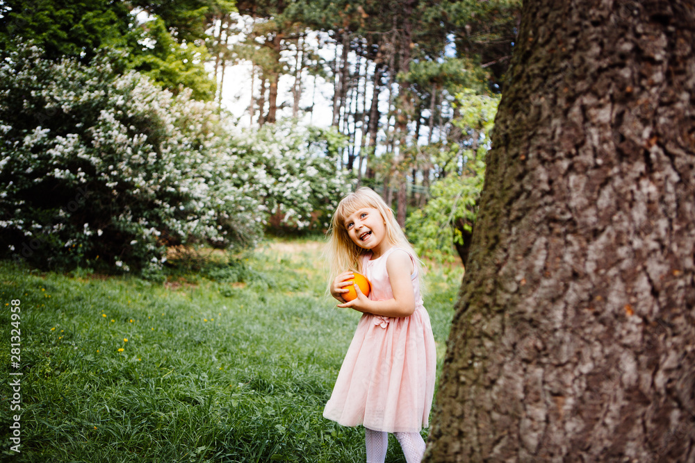 Funny little girl playing with orange in park and smile in summer, childhood and activity
