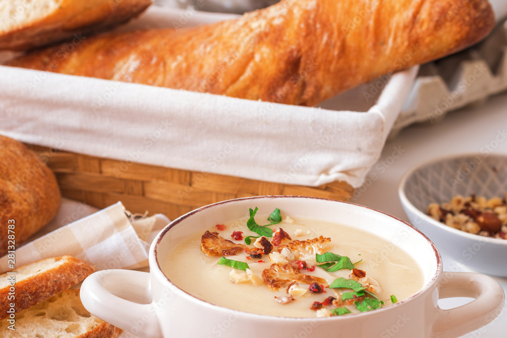 White wooden table with 2 bowls of cauliflower soup embellished with crushed hazelnuts, red pepper, smooth parsley herbs, Swiss twisted bread, frying pan with cauliflower, mortar and yellow flowers. White wooden table with 2 bowls of cauliflower soup embellished with crushed hazelnuts, red pepper, smooth parsley herbs, Swiss twisted bread, frying pan with cauliflower, mortar and yellow flowers.
