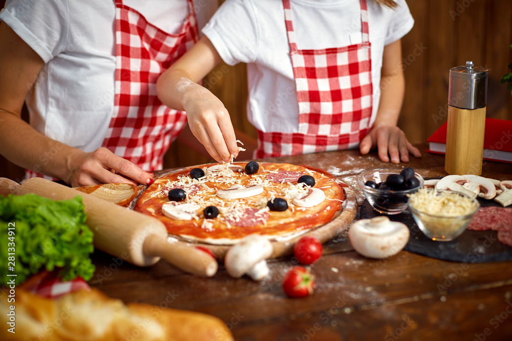 mother and daughter wearing white T-shirts and checkered aprons cooking pizza together and decorating with mushrooms on table filled with ingredients in stylish wooden kitchen