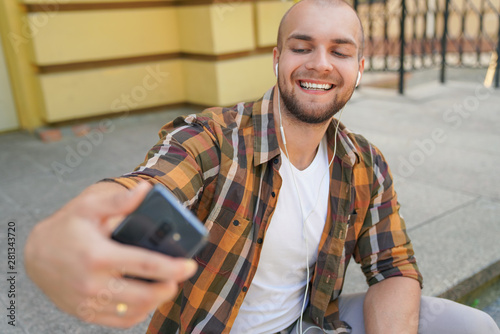 waist up portrait of young good looking bold bearded guy is sitting outdoors on stairs in front of his house making selfie and smiling. he has earphones in his ears. wearing yellow shirt and jeans.