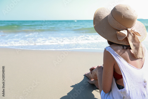 Back view portrait of sexy asian young girl in bikini and hat  looking away to tropical beach while sitting on sandy beach in nature with blue sky.