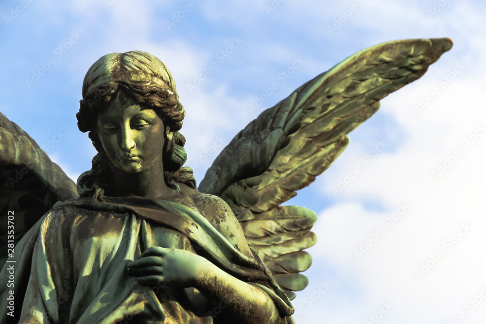 Sad guardian angel bronze sculpture with open long wings across the frame against a bright blue white sky. The sad expression sculpture with eyes down and hand in front of chest.