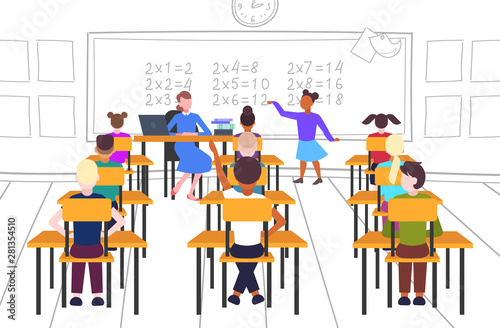 female teacher with pupils sitting at desks looking at schoolgirl solving math problem at chalkboard school lesson education concept modern school classroom interior full length horizontal