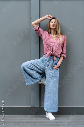 young European girl, natural appearance, long hair, beautiful face, blue wide pants, crimson color shirt, white sneakers cool to pose gray wall background. Linen womens clothing.