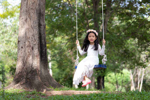 Portrait of little Asian girl playing the swing under the big tree in the nature forest with soft tone processed