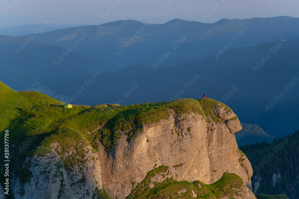 Fototapeta premium Camping on top of the mountain. A person can be seen on the edge of the cliff and her tent installed a few feet away.