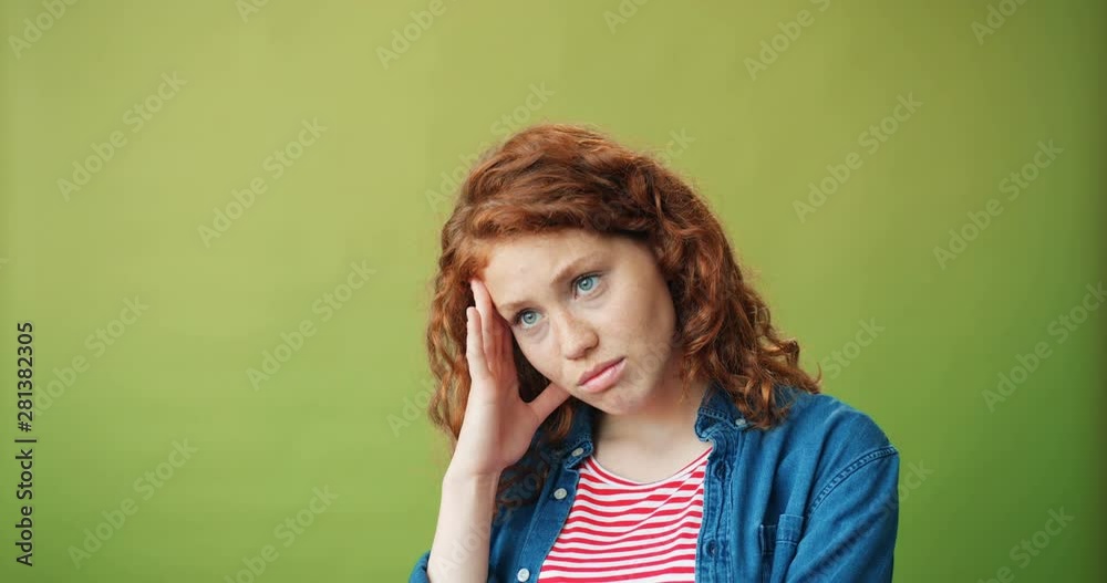 Portrait of unhappy young girl with red hair standing alone touching sad face looking at camera against light green background. Millennials, emotions and sadness concept. Portrait of unhappy young girl with red hair standing alone touching sad face looking at camera against light green background. Millennials, emotions and sadness concept.