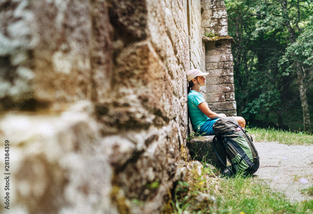 Tired female backpacker resting on the bench near the old antique brick wall castle on the famous Camino de Santiago way.