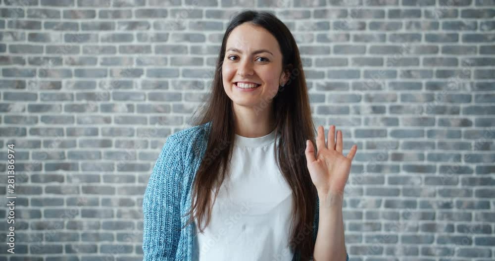 Portrait of friendy woman waving hand and looking at camera with happy face smiling greeting people on brick wall background. Welcome and hello concept. Portrait of friendy woman waving hand and looking at camera with happy face smiling greeting people on brick wall background. Welcome and hello concept.