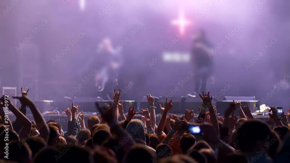 Crowd of people moving rhythmically into the groove with their arms raised, showing horns gesture. Two musicians are performing on the stage. UHD