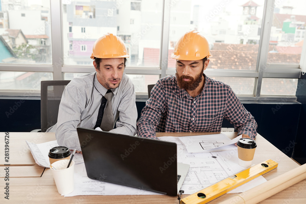 Two business partners in hardhats sitting at the table and working on laptop computer during a meeting at office Two business partners in hardhats sitting at the table and working on laptop computer during a meeting at office