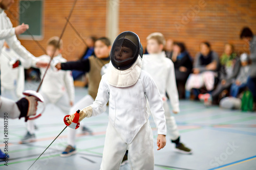 Little kid boy fencing on a fence competition. Child in white fencer uniform with mask and sabre. Active kid training with teacher and children. Healthy sports and leisure. photo