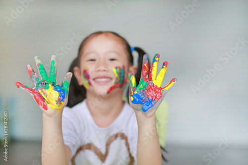 Happy little Asian girl with her colorful hands and cheek painted in the children room. Focus at baby hands.