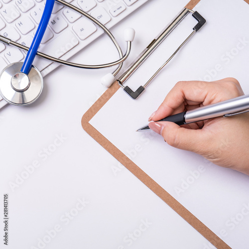 Female doctor writing a medical record case over clipboard on white working table with stethoscope  computer keyboard. Top view  flat lay  copy space