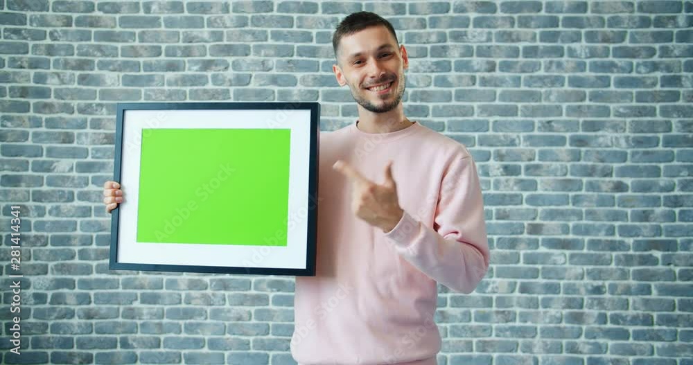 Portrait of handsome man holding chroma key picture frame and showing thumbs-up expressing high evaluation on brick wall background. People and like concept.