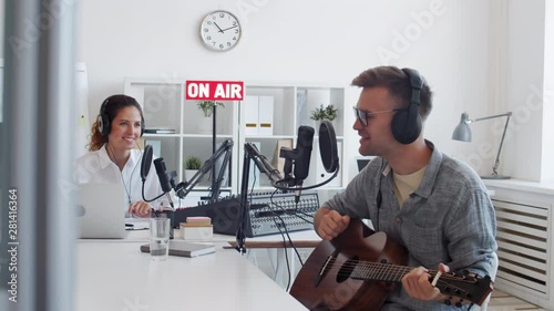 Medium shot of male guest singer sitting at microphone, finishing playing guitar and singing and then female Caucasian radio dj sitting on background starting speaking into mike photo