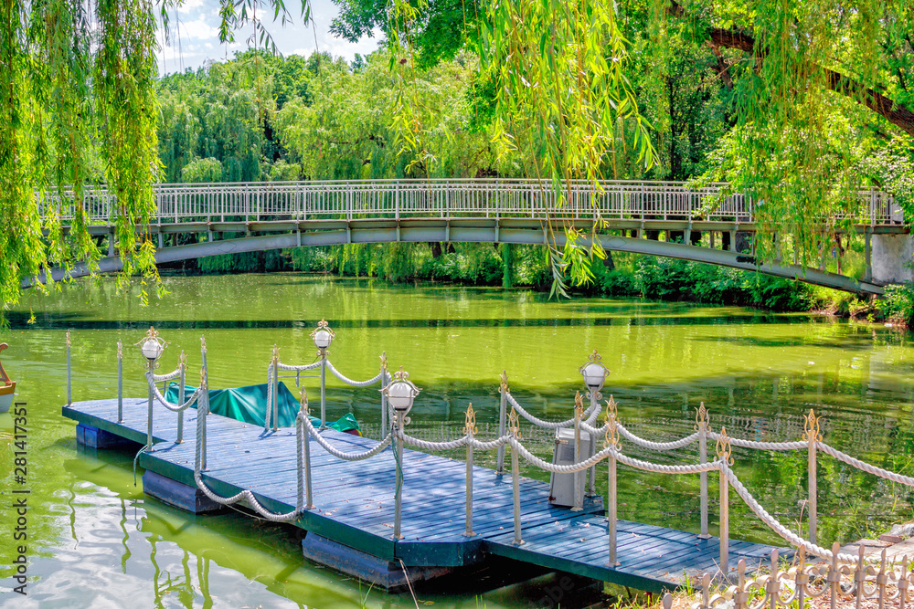 Fototapeta premium View of wooden pier, decorated with lights and rope, in calm green lake water with bridge on background