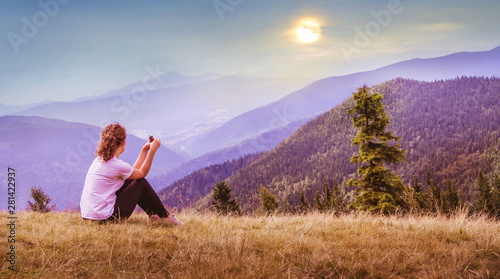 Tableau sur toile A young girl sits on the grass on a background of mountains and watches the sunset