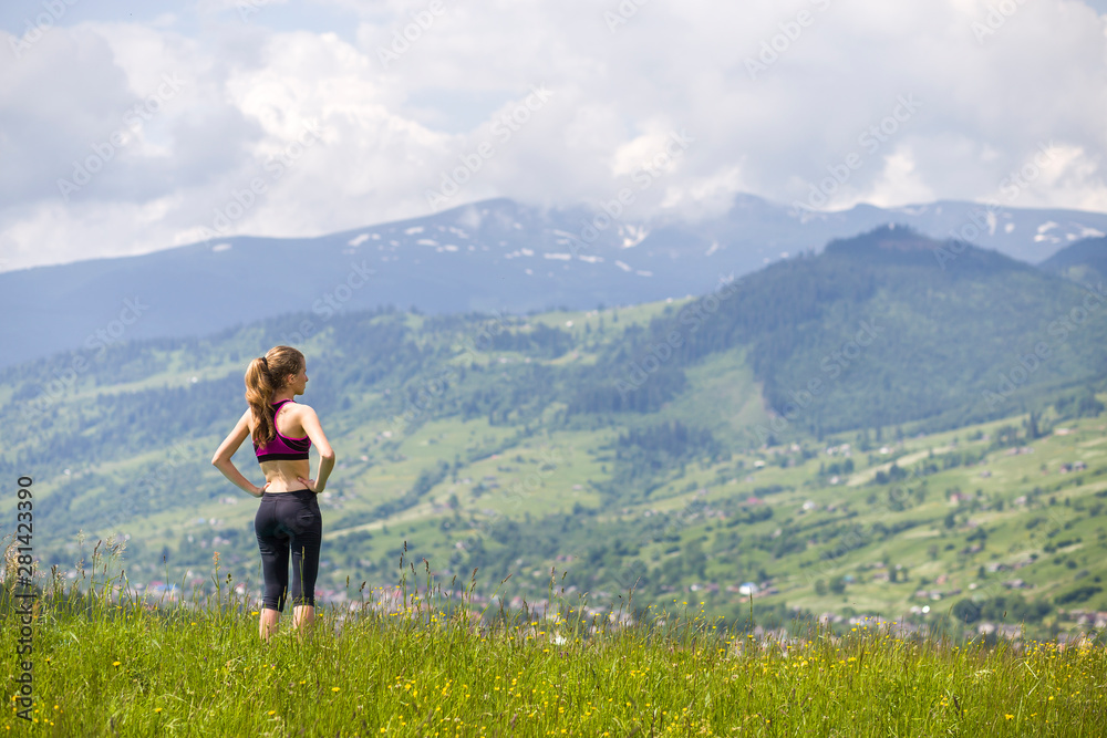 Back view of slim young woman standing on grassy valley on background of green mountains on sunny summer day.