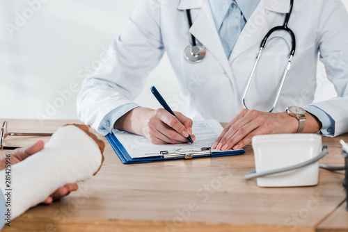 cropped view of patient with broken arm near doctor writing on clipboard