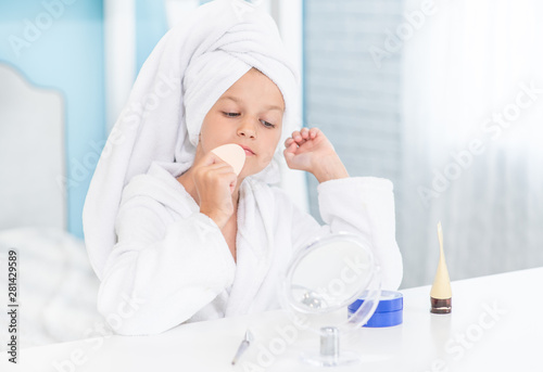 Little girl with a towel on her head puts cream on her face looking in the mirror