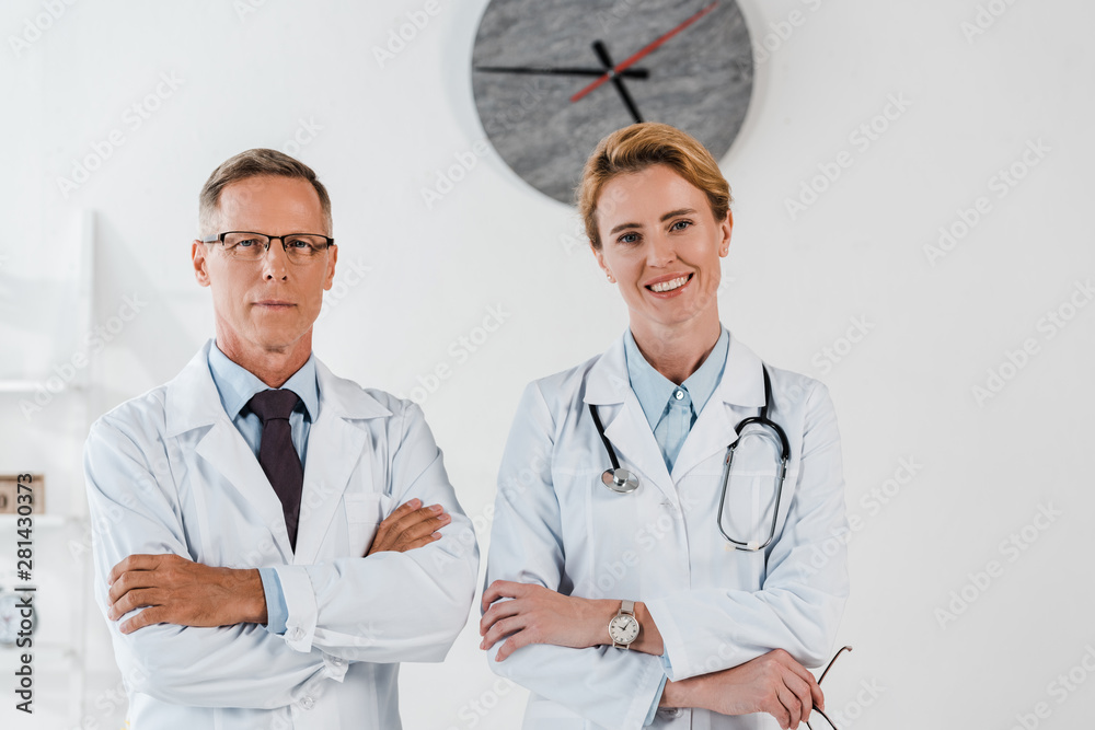 Fototapeta lekarz w okularach stojący ze skrzyżowanymi rękami w pobliżu wesoły kolega w klinice