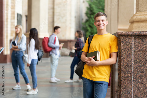 Happy teen guy holding books and smiling in college campus © Prostock-studio