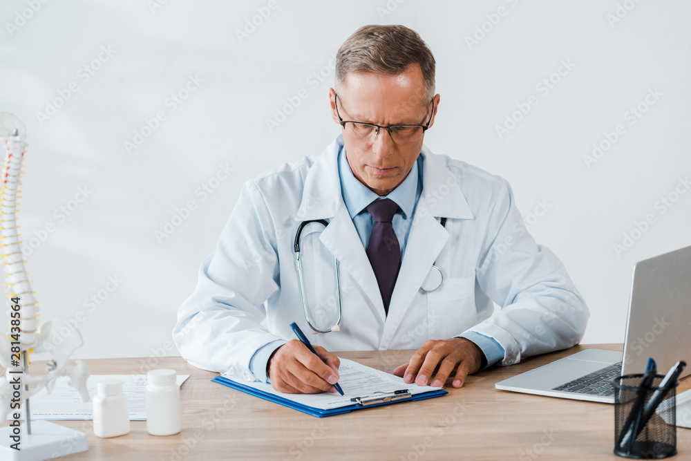 handsome doctor in glasses writing on clipboard near laptop