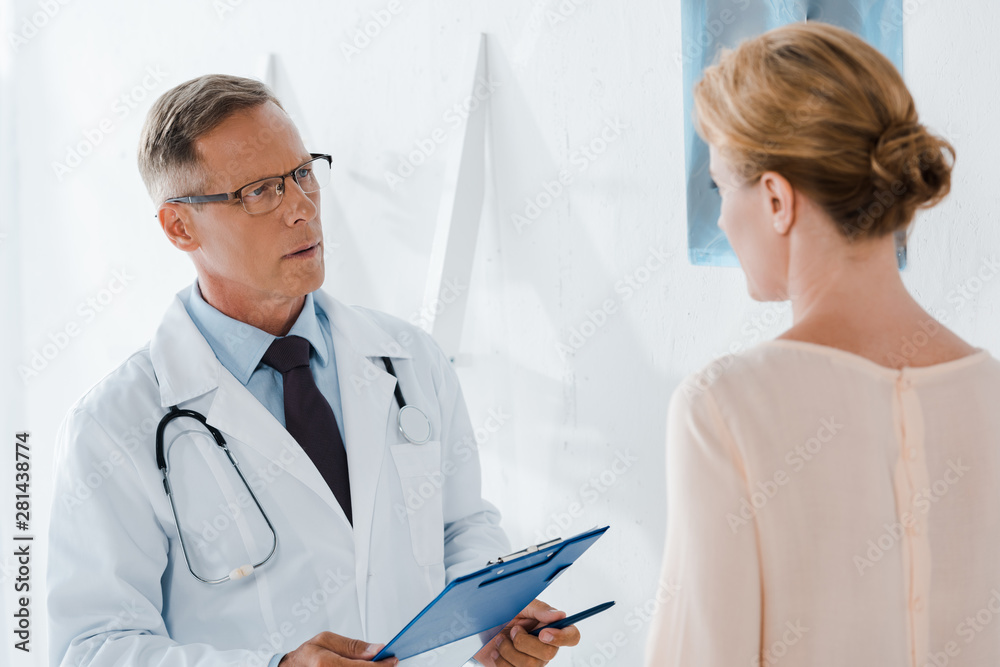 doctor in glasses holding pen and clipboard near patient in clinic doctor in glasses holding pen and clipboard near patient in clinic
