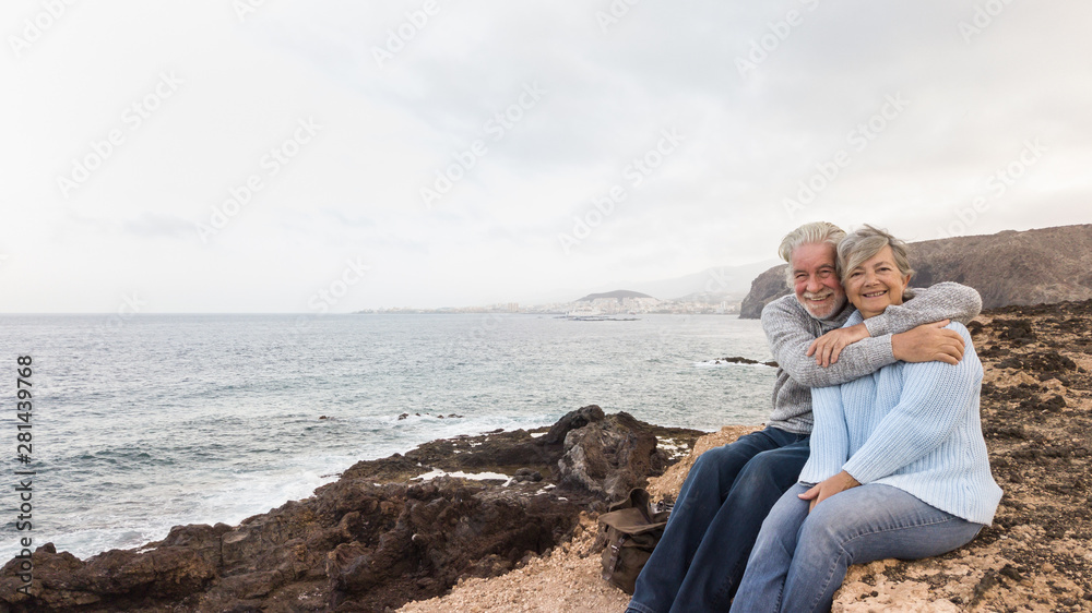Two senior people sitting on the cliff of the ocean and looking at camera Hugging and smiling. Morning soon outside, ready for un healthy excursion. Vacation and happiness Two senior people sitting on the cliff of the ocean and looking at camera Hugging and smiling. Morning soon outside, ready for un healthy excursion. Vacation and happiness