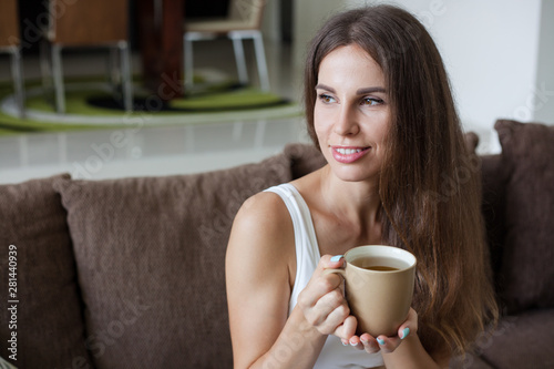 portrait of beautiful smiling brunette girl sitting on sofa in living room and holding mug of tea in her hands. Girl looks away