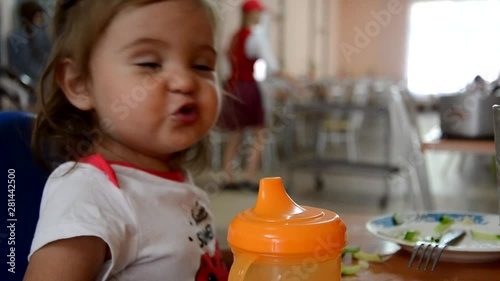 childhood and people concept - little baby drinking from spout cup sitting in highchair at home or at cafe dabbles, disobeys, builds faces, mom tries to feed photo