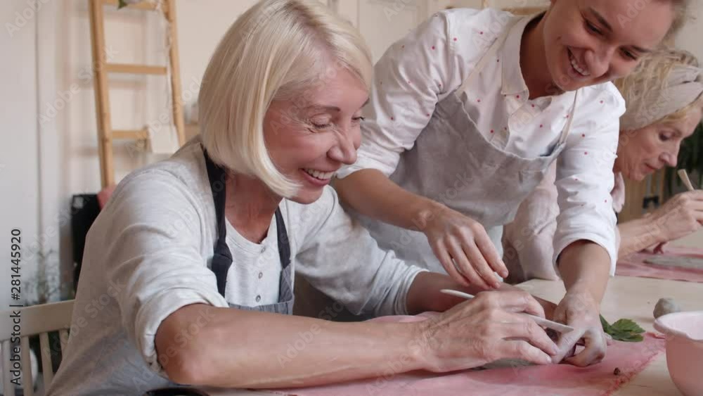 Medium shot of young ceramist standing leaning on worktop and helping middle-aged Caucasian woman sitting nearby decorating flat piece of raw clay Medium shot of young ceramist standing leaning on worktop and helping middle-aged Caucasian woman sitting nearby decorating flat piece of raw clay