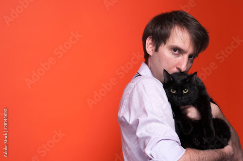 side view of worried man in pink shirt with rolled sleeves holding and hugging cute black cat and looking at camera on coral background with copy space
